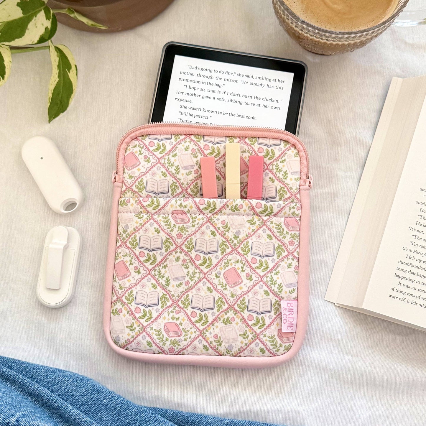 Pink case with small items on a table with a plant, coffee, and book.