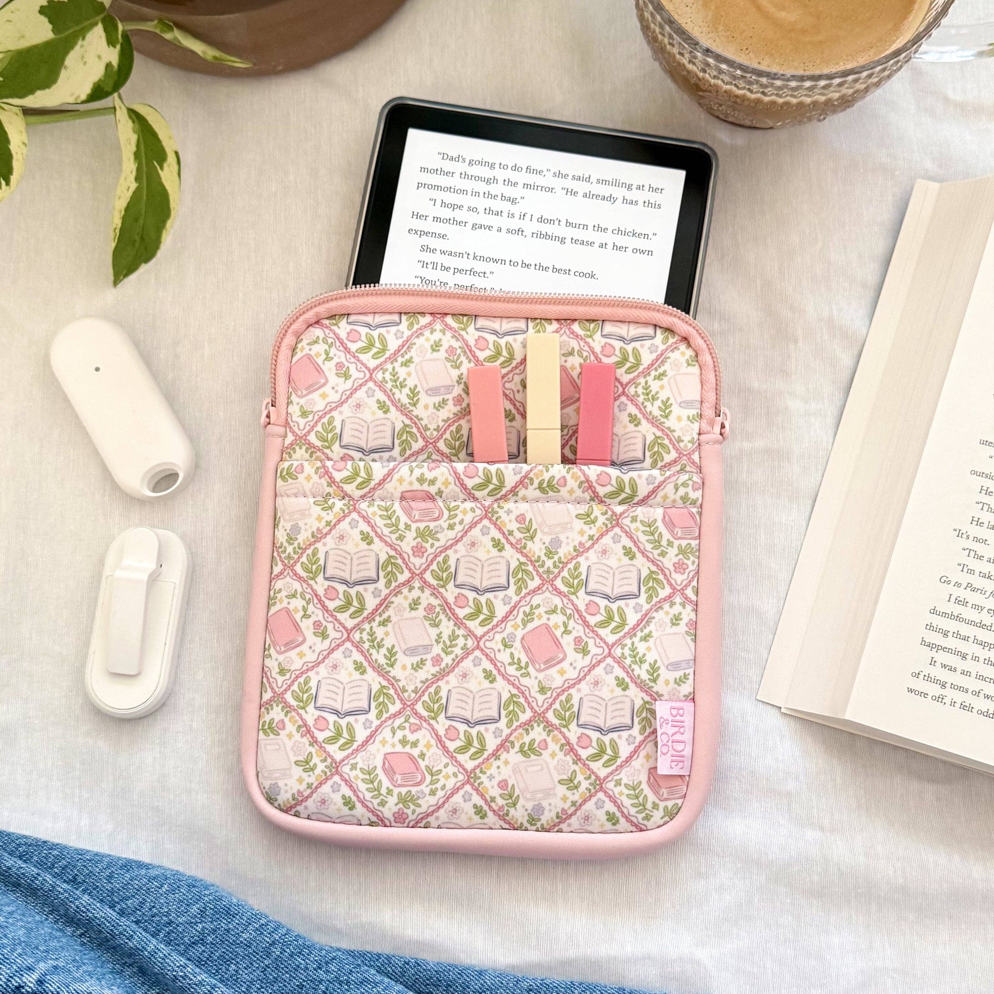 Pink case with small items on a table with a plant, coffee, and book.