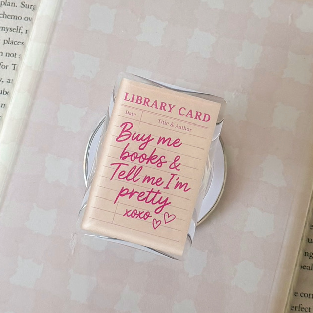 Hand holding a white quilted bag with a pink label over an open book, next to a lit candle and plant.
